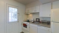 This single-floor residence offers a practical and well-organized layout designed for comfortable everyday living. The kitchen serves as a central highlight, outfitted with white cabinetry that extends to the upper walls, providing ample storage for cookware and pantry essentials. A gas range with an overhead ventilation hood anchors the cooking area, complemented by a countertop sink with a speckled laminate surface that offers functional workspace for meal preparation. The cabinetry and appliances maintain a cohesive, clean aesthetic throughout with a neutral white color palette that brightens the space. A built-in dishwasher adds a layer of convenience to the kitchen setup. Natural light filters in through a window fitted with horizontal blinds, lending an airy feel to the cooking area. A direct exterior door situated adjacent to the stove provides easy access to the outdoors, a practical feature for daily use. The overall ambiance of the space is understated and functional, with light-toned walls and simple finishes that create a clean, welcoming environment suited to a variety of lifestyles.