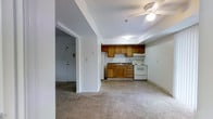 This single-floor space showcases an open-concept design that integrates the kitchen and living area into one cohesive layout. The kitchen is equipped with warm-toned wooden cabinetry, both upper and lower units, providing ample storage. A white electric range and refrigerator offer essential functionality, while a sink with dark countertops completes the workspace. The kitchen transitions seamlessly into the living area, which features neutral-toned carpeting that adds a sense of warmth underfoot, contrasting with the tile flooring in the kitchen zone. A ceiling fan is centrally mounted, providing both lighting and air circulation throughout the open space. The walls are finished in a clean, neutral white, contributing to a bright and airy ambiance. A doorway visible from the main area leads to an entry vestibule, where a solid entry door with a standard lock provides access to the unit. The overall layout is practical and efficient, making good use of the available space while maintaining an open, uncluttered feel suitable for everyday living.