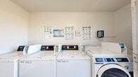 This shared laundry facility is a single-room space designed for efficient, communal use. The room is finished with light-toned luxury vinyl plank flooring and neutral white walls, creating a clean and bright environment. Along one wall, a row of top-loading and front-loading washers — branded by Maytag and operating through the WASH IBO 470 payment system — are neatly arranged side by side. These coin-operated machines are clearly labeled and accompanied by prominently posted instructional guides covering machine operation and laundry best practices, ensuring ease of use for all residents. On the adjacent wall, stacked commercial dryers by WASH provide additional capacity, maximizing the use of the available space. A wall-mounted paper towel dispenser adds a practical touch. The room is accessed through a distinctive light blue door fitted with a security grate, allowing natural light and outdoor views to filter in while maintaining safety. A fire extinguisher cabinet is mounted near the entrance, reflecting attention to safety standards. The overall layout is straightforward and functional, making efficient use of the compact footprint while providing all essential laundry amenities in a tidy, well-maintained setting.