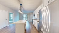 Located in Downers Grove, IL, this modern single-floor residence offers a thoughtfully designed open-concept layout with clean, contemporary finishes throughout. The kitchen serves as the centerpiece of the living area, featuring crisp white shaker cabinetry with matte black hardware, a large center island with a quartz countertop, subway tile backsplash, and a full suite of stainless steel appliances including a gas range, over-the-range microwave, dishwasher, and refrigerator. A stylish multi-pendant light fixture illuminates the island, adding a sleek, modern touch. Light wood-tone flooring flows seamlessly through the main living areas, complementing the soft, neutral wall tones. The living area opens to a private balcony through sliding glass doors, allowing natural light to fill the space and providing views of the surrounding building exterior. Bedrooms are carpeted for comfort and feature large windows that bring in additional natural light. The bathroom is well-appointed with a white vanity, quartz countertop, a large mirror, and wood-look flooring. Clean lines, neutral tones, and modern fixtures combine to create a cohesive, move-in-ready living environment throughout the entire residence.