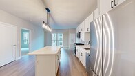 Located in Downers Grove, IL, this modern single-floor residence offers a thoughtfully designed open-concept layout with clean, contemporary finishes throughout. The kitchen serves as the centerpiece of the living area, featuring crisp white shaker cabinetry with matte black hardware, a large center island with a quartz countertop, subway tile backsplash, and a full suite of stainless steel appliances including a gas range, over-the-range microwave, dishwasher, and refrigerator. A stylish multi-pendant light fixture illuminates the island, adding a sleek, modern touch. Light wood-tone flooring flows seamlessly through the main living areas, complementing the soft, neutral wall tones. The living area opens to a private balcony through sliding glass doors, allowing natural light to fill the space and providing views of the surrounding building exterior. Bedrooms are carpeted for comfort and feature large windows that bring in additional natural light. The bathroom is well-appointed with a white vanity, quartz countertop, a large mirror, and wood-look flooring. Clean lines, neutral tones, and modern fixtures combine to create a cohesive, move-in-ready living environment throughout the entire residence.