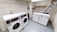 This single-floor laundry room is a dedicated, utilitarian space designed to meet practical laundry needs. The room features multiple commercial-grade front-loading washing machines and top-loading dryers, all finished in white, arranged along the walls to maximize usable floor space. The machines appear to be coin-operated, suggesting shared or common-use functionality.

The room has a straightforward layout with painted concrete or drywall surfaces and a smooth, painted concrete floor in a light grey tone. Overhead fluorescent lighting provides bright, even illumination throughout the space. Exposed utility piping is visible along the upper walls, indicating direct plumbing connections to the machines. An electrical panel is mounted on one wall, along with visible conduit and wiring that support the room's operational infrastructure.

The overall aesthetic is purely functional, with no decorative elements. The space is compact but efficiently organized, allowing users to move between machines with ease. The white walls and bright lighting contribute to a clean, well-lit environment suited to its purpose as a shared laundry facility.