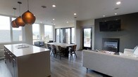 This single-floor space showcases a thoughtfully designed open-concept layout that seamlessly blends living, dining, and entertaining areas. The interior is finished with wide-plank hardwood-style flooring in a cool gray tone, complementing the clean, neutral walls and contemporary aesthetic throughout.

The dining area serves as a central focal point, anchored by a substantial rustic wood dining table surrounded by dark wood chairs — a striking contrast that adds warmth and character to the modern setting. Overhead, recessed lighting provides even, ambient illumination across the space.

The living area features a sleek sofa and is anchored by a wall-mounted flat-screen television above a modern gas fireplace set against a dark accent wall, creating a cozy yet sophisticated atmosphere.

Large floor-to-ceiling windows and a set of French doors flood the space with natural light and open directly onto an outdoor terrace, offering panoramic views of the surrounding cityscape and landscape. A secondary door provides additional outdoor access, reinforcing the strong connection between the interior and exterior living spaces.

A pendant light fixture with a warm copper tone adds a refined decorative touch, tying together the space's blend of rustic and contemporary design elements. Overall, the space exudes a bright, welcoming, and stylish ambiance ideal for both everyday living and entertaining.