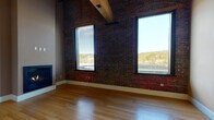 This single-floor unit showcases a compelling blend of industrial character and modern finishes. The open living area immediately impresses with soaring ceilings supported by exposed timber beams, original brick walls, and gleaming hardwood flooring throughout. Large black-framed windows flood the space with natural light while framing picturesque views of the surrounding landscape and treeline. A built-in gas fireplace serves as a focal point in the main living space, adding warmth and ambiance.

The kitchen is well-appointed with rich cherry wood cabinetry, granite countertops, stainless steel appliances including a range, microwave, and refrigerator, and continued hardwood flooring. Exposed structural columns add to the loft-like industrial aesthetic of the kitchen and living areas.

The bedrooms feature plush carpeting, tall brick accent walls, exposed timber beams, and generous windows that maintain the bright, airy feel found throughout the unit. The bathroom is finished with a granite-topped vanity with cherry cabinetry, tile flooring, a shower-tub combination, and the added convenience of an in-unit stacked washer and dryer. Together, these elements create a space that is both functional and rich in architectural character.