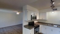 This single-floor space is designed with a practical, open layout that connects the kitchen and living areas seamlessly. The kitchen is well-appointed with white cabinetry extending to the ceiling, providing ample storage. Dark grey countertops offer a subtle contrast to the white cabinets, while a black electric range and stainless steel microwave supply essential cooking functionality. A sink is positioned along the counter, and track lighting above adds focused illumination to the workspace.

The adjoining living area continues the dark hardwood-style flooring found throughout, contributing to a cohesive aesthetic across the space. A ceiling fan with a light fixture serves the living area, providing both air circulation and ambient lighting. Neutral white walls keep the interior feeling bright and clean, while a textured popcorn ceiling is consistent throughout. A hallway leads from the main living area toward additional rooms, hinting at further functional spaces beyond. The overall ambiance is clean, straightforward, and move-in ready, suited to those seeking a practical and low-maintenance living environment.