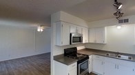 This single-floor space is designed with a practical, open layout that connects the kitchen and living areas seamlessly. The kitchen is well-appointed with white cabinetry extending to the ceiling, providing ample storage. Dark grey countertops offer a subtle contrast to the white cabinets, while a black electric range and stainless steel microwave supply essential cooking functionality. A sink is positioned along the counter, and track lighting above adds focused illumination to the workspace.

The adjoining living area continues the dark hardwood-style flooring found throughout, contributing to a cohesive aesthetic across the space. A ceiling fan with a light fixture serves the living area, providing both air circulation and ambient lighting. Neutral white walls keep the interior feeling bright and clean, while a textured popcorn ceiling is consistent throughout. A hallway leads from the main living area toward additional rooms, hinting at further functional spaces beyond. The overall ambiance is clean, straightforward, and move-in ready, suited to those seeking a practical and low-maintenance living environment.