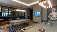This sophisticated single-floor entertainment and game lounge is thoughtfully designed for social gatherings and leisure. The centerpiece of the space is a pair of full-size billiard tables with rustic wood frames, complemented by a striking geometric patterned area rug that anchors the room with visual depth. Overhead, elegant crystal pendant chandeliers and recessed cove lighting cast a warm, refined ambiance throughout. A large wall-mounted flat-screen television is seamlessly integrated into a structured wall column, making the space equally suited for watching events or sports. A cushioned lounge banquette with throw pillows runs along one wall, offering comfortable seating adjacent to a large decorative mirror that amplifies the sense of space. The warm-toned walls, tray ceiling details, and wood-plank flooring transitions contribute to an inviting yet polished aesthetic. The overall design balances a modern upscale sensibility with casual, recreational functionality, making it an ideal venue for community entertainment, social events, or friendly competition.