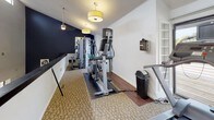 The Abbington Hills Fitness Center is a thoughtfully designed exercise space that combines functionality with a clean, modern aesthetic. The facility features notably high ceilings with a row of clerestory windows running along the upper wall, allowing abundant natural light to fill the room and contributing to the open, spacious atmosphere. Ceiling fans with pendant lighting fixtures provide both air circulation and warm supplemental illumination throughout the space.

The workout area is equipped with a variety of cardio machines, including ellipticals, treadmills, and stair climbers, arranged along the carpeted floor to maximize usability. Rubber exercise mats are positioned beneath select equipment for added stability and floor protection. A dedicated strength training station is also available, offering a well-rounded fitness experience.

The interior design features a crisp white and deep navy color scheme, with one boldly painted accent wall adding visual contrast. A collection of colorful abstract artworks is displayed on a half-wall partition, lending a creative touch to the environment. The combination of natural light, high ceilings, and tasteful décor creates an inviting atmosphere that encourages an active and comfortable workout experience.