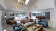 The Enchanted Clubhouse is a thoughtfully designed single-story communal space that combines comfort and style in an open, welcoming layout. The interior is anchored by striking vaulted ceilings that amplify the sense of spaciousness, while multiple skylights flood the space with abundant natural light throughout the day. Elegant globe pendant lights hang at varying heights, complementing the architectural design and adding a refined touch to the atmosphere.

The lounge area features a neutral-toned sofa accented with warm terracotta throw pillows, paired with blue upholstered armchairs and a rustic wooden coffee table, creating an inviting and relaxed seating arrangement. Decorative elements such as a sculptural orb accent and curated tabletop accessories add personality to the space.

Adjacent to the lounge, a dedicated dining area with orange upholstered chairs and a long table is set against a cool blue accent wall, providing a distinct and functional zone for gatherings. Large windows and sliding glass doors frame views of the outdoor pool area, seamlessly connecting the interior to the exterior and enhancing the bright, airy ambiance throughout.