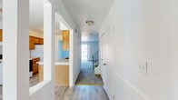 This two-story residence offers a practical and well-organized layout across both levels. The main floor welcomes you with a central hallway finished in wood-look flooring that transitions smoothly into carpeted living areas. The galley-style kitchen is outfitted with warm oak cabinetry, light countertops, a coil-top electric range, a refrigerator, a dishwasher, and a double-basin sink — all oriented toward a doorway that opens directly to the living area and beyond to an outdoor space. The spacious living area is carpeted and bathed in natural light through a large sliding glass door fitted with vertical blinds, providing access to an exterior patio or yard area. A staircase with a metal railing connects the main floor to the upper level. Upstairs, carpeted bedrooms offer a quiet retreat, each featuring louvered closet doors for ample storage. The bathrooms are finished with wood-toned vanity cabinetry, light countertops, and wood-look tile flooring. One bathroom includes a tub-shower combination and a distinctive octagonal window that frames a view of surrounding greenery, adding a charming architectural detail. Throughout, the home maintains a clean, neutral palette with white walls that lend a bright, airy feel to every room.
