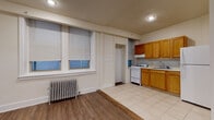 This single-floor unit offers a practical and straightforward layout across several rooms, reflecting classic urban residential architecture. The living and bedroom areas feature warm hardwood-style flooring and neutral walls, creating a clean, versatile canvas. Each room is equipped with cast-iron radiators, a hallmark of traditional building construction, ensuring consistent heating throughout. Multiple windows fitted with horizontal blinds allow natural light to filter into the rooms while maintaining privacy, with street-level views adding a sense of connectivity to the surrounding neighborhood.

The kitchen is outfitted with warm-toned wooden cabinetry, a white gas range, a full-size refrigerator, and a single-basin sink, providing all essential cooking amenities. Light ceramic tile flooring in the kitchen offers a practical contrast to the wood flooring in the living spaces. A separate doorway leads to an additional room or storage area adjacent to the kitchen.

The bathroom features classic white subway tile walling, a tiled floor, a window with blinds for ventilation and light, and a cast-iron radiator consistent with the rest of the unit. The overall layout is efficient and functional, with clearly defined spaces suitable for comfortable everyday living.