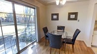 This single-floor residence offers a well-organized layout with distinct living areas, each thoughtfully designed for everyday comfort. The dining area is a standout space, anchored by a round glass-top table surrounded by dark leather chairs. A large picture window floods the room with natural light and frames a lush, tree-lined outdoor view, while a ceiling fan with a light fixture overhead adds a functional touch. Framed artwork adorns the walls, completing the inviting atmosphere.

The living room features plush carpeting and a spacious sectional sofa, creating a relaxed setting for lounging. An open entryway connects to the foyer, where hardwood flooring, a round decorative mirror, and a potted plant add warmth to the transition space. A media console with storage anchors the living area.

The galley-style kitchen is efficiently designed with white cabinetry lining both sides, offering ample storage. Tile flooring, a gas range, a refrigerator, and a butcher block countertop contribute to its practicality. A built-in pantry with open shelving provides additional storage.

A shared building entryway features patterned wallpaper, carpeted stairs with a wrought iron railing, and an arched window that draws in natural light, lending a classic character to the common area.