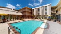 This communal pool area is situated within a multi-building complex in Metairie, Louisiana, designed around a central courtyard that promotes an open, social atmosphere. The centerpiece is a generously sized rectangular swimming pool with clear, blue water, surrounded by a tiled pool deck with lounge chairs and yellow shade umbrellas, ideal for relaxing outdoors under Louisiana's sunny skies.

The pool is enclosed by a black wrought-iron security fence with a gated entry, ensuring both safety and privacy for residents. The surrounding buildings feature a mid-century architectural style, with brick and textured facades, covered balconies, and large windows that overlook the courtyard. A covered walkway runs along the perimeter of the complex, providing shaded passage between areas.

The common corridor leading to the pool area houses a substantial bank of individual U.S. Mail mailboxes mounted along the wall, offering convenient mail access for all residents. This covered hallway opens toward the parking area and exterior, keeping foot traffic organized and sheltered. Together, these shared amenities create a functional and comfortable community environment well-suited to the warm climate of the greater New Orleans area.