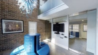 This distinctive single-floor space makes an immediate impression through its dramatic architectural contrasts and carefully curated design elements. Exposed brick walls in warm amber tones provide a raw, industrial backdrop that is artfully balanced by crisp white structural columns and modern finishes throughout.

The living area commands attention with soaring double-height ceilings, creating an expansive, open atmosphere. A mezzanine-level overhang with glass railings adds visual depth and architectural interest, allowing light to flow freely through the vertical space. A wall-mounted flat-screen television is neatly integrated into a white media column, with dedicated shelving for media components beneath.

Bold blue tufted leather seating anchors the main living zone, providing a vibrant focal point that complements the industrial aesthetic. Overhead, an ornate blue blown-glass chandelier adds an artistic, sculptural element that ties the color palette together cohesively.

Large plantation shutters on the windows offer precise light control while contributing to the clean, refined character of the interior. Glass partition walls and sliding doors define adjacent spaces without sacrificing openness, maintaining visual continuity throughout. The light-toned tile flooring grounds the space and reflects natural light effectively.