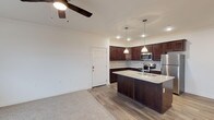 The BLVD At Hays offers a thoughtfully designed single-floor layout that seamlessly blends comfort and modern style. The open-concept living and kitchen area serves as the heart of the space, creating an inviting and functional environment. The kitchen is a standout feature, showcasing rich dark cabinetry that provides generous storage, complemented by sleek granite countertops and a peninsula island with an undermount sink — ideal for both meal preparation and casual dining. Pendant lighting hangs above the island, adding a warm, contemporary touch, while recessed lighting throughout ensures the space is well-lit. Stainless steel appliances, including a refrigerator, microwave, and range, complete the modern kitchen aesthetic. The living area flows naturally from the kitchen, featuring durable wood-style flooring that transitions into plush carpeting, offering a comfortable and versatile space. A ceiling fan anchors the room, providing both functionality and a finishing design detail. Clean white walls and crisp trim maintain a bright, neutral palette throughout, making the space feel open and move-in ready. The unit's single-floor design ensures easy accessibility and a cohesive flow from one area to the next.