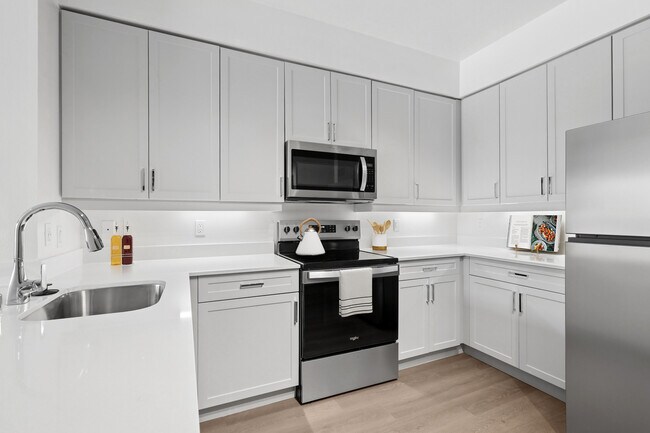 Cocina renovada del Paquete II con electrodomésticos de acero inoxidable, gabinetes grises, encimeras de cuarzo blanco y pisos de superficie dura. - Avalon at Cahill Park