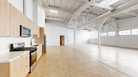 Located in the vibrant South Los Angeles corridor at 2348 S Santa Fe Ave, this single-floor industrial loft offers a generous and highly versatile open-plan layout. The space is defined by its dramatic soaring ceilings with exposed, white-painted structural trusses and large-scale HVAC ductwork that lend an authentic warehouse aesthetic. Polished concrete floors run throughout, tying the entire space together with a clean, industrial character.

Natural light pours in through rows of oversized industrial-style grid windows along the upper walls, illuminating the bright white interior and creating an airy, open ambiance. The main open area is expansive, offering exceptional flexibility for a variety of uses.

A compact kitchen is integrated along one wall, featuring light wood cabinetry, stainless steel appliances including a refrigerator and range, and a sink — providing functional amenities within the open floor plan. A dedicated utility area includes a utility sink and additional storage. The full bathroom is thoughtfully finished with a light wood vanity, a framed mirror with matte black fixtures, and a tiled soaking tub with shower combination. Skylights in the ceiling supplement the window light, ensuring the space remains bright throughout the day.