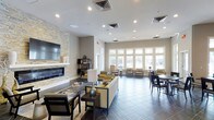 This single-floor communal space is thoughtfully designed to accommodate a variety of social and casual activities. The open-plan layout is anchored by a striking stacked-stone accent wall that frames a sleek linear fireplace, above which a large flat-screen television is mounted — creating a warm and inviting focal point for the lounge area. Comfortable seating arrangements with neutral-toned sofas and accent chairs in gold and grey are positioned to encourage conversation and relaxation.

Large, multi-pane windows line the exterior walls, flooding the interior with generous natural light and offering views of the surrounding outdoor landscape. The expansive tile flooring in a slate-grey finish extends throughout, tying the space together cohesively.

A bar-height counter with tall leather chairs provides a dedicated social dining area, complemented by pendant lighting in an industrial cage style that adds a modern touch. Additional dining tables and chairs are arranged nearby for flexible seating options. Recessed ceiling lighting throughout ensures the space remains well-lit, while the neutral warm-beige walls contribute to the overall contemporary and welcoming ambiance.