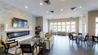 This single-floor communal space is thoughtfully designed to accommodate a variety of social and casual activities. The open-plan layout is anchored by a striking stacked-stone accent wall that frames a sleek linear fireplace, above which a large flat-screen television is mounted — creating a warm and inviting focal point for the lounge area. Comfortable seating arrangements with neutral-toned sofas and accent chairs in gold and grey are positioned to encourage conversation and relaxation.

Large, multi-pane windows line the exterior walls, flooding the interior with generous natural light and offering views of the surrounding outdoor landscape. The expansive tile flooring in a slate-grey finish extends throughout, tying the space together cohesively.

A bar-height counter with tall leather chairs provides a dedicated social dining area, complemented by pendant lighting in an industrial cage style that adds a modern touch. Additional dining tables and chairs are arranged nearby for flexible seating options. Recessed ceiling lighting throughout ensures the space remains well-lit, while the neutral warm-beige walls contribute to the overall contemporary and welcoming ambiance.