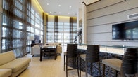 This refined single-floor space makes an immediate impression with its dramatic double-height ceilings and expansive floor-to-ceiling windows dressed in sheer, flowing curtains that diffuse natural light throughout the interior. The open-plan layout seamlessly connects a comfortable lounge area — furnished with neutral-toned sofas and a dining table with dark wood chairs — to a sleek bar or counter space finished in a striking mosaic tile facade. The bar features a glass countertop and dark bar stools, creating an inviting spot for casual seating or entertaining. Light hardwood flooring runs throughout, complementing the warm, recessed ceiling lighting that adds a soft ambient glow. Tall decorative columns and clean white walls contribute to the space's modern, upscale aesthetic, while carefully placed sculptural accents and lush potted plants add touches of organic warmth. The generous glazing along the exterior wall connects the interior to the outdoors, filling the space with a sense of openness and brightness. Overall, the space presents a polished, contemporary environment well-suited for social gatherings, relaxation, or professional use.