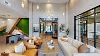 The Evolve at the Pines clubhouse at 1215 Evolve Way in Greenville, NC, is a thoughtfully designed community amenity space that blends contemporary style with functional comfort across a single, generously sized floor. The grand entrance lounge immediately sets the tone with soaring double-height ceilings, herringbone hardwood flooring, and a striking living moss wall accent alongside warm pendant lighting. Curated seating areas feature leather and neutral upholstered furniture arranged around a central coffee table, creating an inviting social hub. A dramatic floor-to-ceiling steel-framed glass partition separates the lounge from an interior conference and gathering room, maintaining openness while defining distinct zones. The main living area anchors around a bold dark tile fireplace wall with an integrated linear fireplace, a wall-mounted TV, and oversized brass drum pendant fixtures that add a luxurious finish. The kitchen and social bar area is equally impressive, showcasing a long stone-topped island with bar seating, bold olive-green cabinetry, geometric pendant lights, a shimmering mosaic tile backsplash, and artistic wall murals. A dedicated co-working office features a built-in desk, teal velvet seating, an arched accent wall, and large black-framed windows that frame lush outdoor greenery, providing an inspiring and productive workspace.