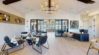 This thoughtfully designed single-story community clubhouse blends modern farmhouse aesthetics with contemporary comfort throughout its well-appointed spaces. The grand entrance lobby greets visitors with soaring vaulted ceilings, exposed dark wood beams, and an elegant chandelier, all set above light wood-look tile flooring. Floor-to-ceiling glass doors with sleek black steel framing connect the main lounge areas, flooding the interiors with natural light and offering views of the outdoor pool area.

The main lounge features a striking white paneled fireplace wall with curated artwork, comfortable seating arrangements, and a large entertainment wall with a mounted flat-screen TV framed in rich wood paneling. A separate game and social lounge offers a full shuffleboard table, a dark island with bar seating, and sliding glass access to the pool courtyard, all beneath vaulted ceilings with an industrial pendant light fixture.

A private conference room provides an intimate setting with exposed brick walls, tall windows with sheer drapery, leather seating, and a wall-mounted display screen — ideal for meetings or focused work sessions.

The expansive fitness center is equipped with treadmills, ellipticals, cable machines, multi-functional strength rigs, and plyometric boxes, all bathed in natural light from large windows with exterior views. The clubhouse as a whole delivers a polished, resort-inspired atmosphere designed for community living.