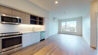 This contemporary single-floor residence showcases a clean, modern design with warm hardwood-style flooring that flows seamlessly throughout the entire space. The open-concept layout connects the kitchen and living area, creating a bright and functional environment. The kitchen is well-appointed with stainless steel appliances—including a refrigerator, range, microwave, and dishwasher—complemented by a mix of white flat-panel cabinetry and natural wood open shelving with under-cabinet lighting. A wall-mounted mini-split unit provides climate control to the living area, which benefits from a large picture window offering expansive city skyline views and abundant natural light. The bedroom is a serene, minimalist space featuring a tall, narrow window that frames additional urban views while maintaining privacy. The full bathroom is finished in a contemporary style, featuring a floating white vanity with a large mirror, a tiled tub-shower combination, and consistent hardwood-style flooring. The overall ambiance of the unit is light, airy, and thoughtfully designed, making it a polished urban residence with a cohesive aesthetic throughout.