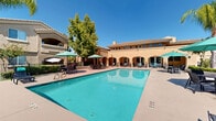 Located at 1601 Vineyard Rd in Roseville, California, this outdoor amenity area showcases a resort-style design inspired by Mediterranean architecture. The centerpiece is a generously sized swimming pool with crystal-clear turquoise water, flanked by a broad sundeck furnished with lounge chairs and teal-shaded umbrellas, ideal for relaxing under the California sun. Adjacent to the main pool, a separate circular spa offers a more intimate retreat, surrounded by manicured hedgerows and lush tropical plantings including towering palm trees and mature shade trees. The surrounding building features warm stucco exteriors in earthy gold tones, accented by arched colonnades and a covered loggia that provides shaded seating and dining areas directly overlooking the pool. Wrought-iron fencing borders portions of the space, adding a refined, classic touch while maintaining open sightlines to the verdant landscaping beyond. Potted plants and colorful ornamental grasses add layers of visual interest throughout the grounds. The overall ambiance is bright, open, and welcoming, with abundant natural light and a well-maintained, cohesive design that creates an inviting outdoor living environment perfectly suited to the warm Roseville climate.