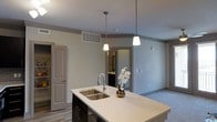 This single-floor residence showcases a clean, modern aesthetic throughout its open-concept layout. The kitchen serves as a central highlight, anchored by a large island topped with a crisp white quartz countertop and an undermount double-basin stainless steel sink paired with a sleek pull-down faucet. Dark cabinetry on the island provides a striking contrast against the light countertop and neutral-toned walls, lending the space a contemporary character. Two cone-shaped pendant lights hang above the island, providing focused task lighting while enhancing the overall design. The open floor plan connects the kitchen seamlessly to the adjacent living area, creating a sense of spaciousness and ease of flow between spaces. Light wood-look flooring runs throughout, adding warmth to the neutral palette of soft gray walls and white ceilings. A glimpse into an adjoining room reveals additional cabinetry and fixtures, suggesting a well-appointed bathroom or utility space nearby. A paneled door off the kitchen leads to what appears to be a storage or pantry area, adding functional depth to the layout. The space is illuminated by a combination of natural and recessed lighting, creating a bright and welcoming ambiance throughout.