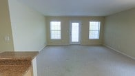 This single-floor space offers a clean, functional layout designed for comfortable everyday living. The main living area is generously sized, featuring wall-to-wall carpeting in a neutral tone that adds warmth underfoot and complements the soft beige walls throughout. Three large windows — including two flanking a glass-paneled entry door — allow abundant natural light to flow into the space, creating a bright and welcoming atmosphere. The entry door itself features a full-length glass panel, further enhancing the connection between the interior and the outside. White baseboards frame the room neatly, contributing to a crisp, well-maintained appearance. A partial view of an adjacent kitchen area reveals light wood cabinetry and a stone-look countertop, suggesting a practical and well-appointed kitchen space nearby. The open feel of the main living area provides flexibility for a variety of furniture arrangements and uses. The overall ambiance is calm and neutral, offering a versatile blank canvas ready to be personalized. The space is well-suited for those seeking a straightforward, move-in ready environment with ample natural light and a cohesive, understated aesthetic throughout.