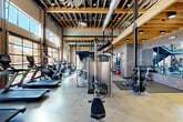 Located at 10720 Linkwood Ct in Baton Rouge, LA, this two-floor fitness and recreation facility blends industrial design with rustic warmth across a generously sized footprint. The main floor serves as the heart of the space, housing a comprehensive gym area with an impressive array of cardio machines — including treadmills and stair climbers — alongside cable machines, free weights, barbells, and dedicated stretching zones. Soaring exposed wood beam ceilings, raw brick accent walls, and polished concrete floors give the gym a distinctive industrial-rustic character, while oversized glass garage doors flood the space with natural light and provide open views of the outdoors. A dedicated group fitness studio on the main level features wood plank flooring, brick walls, and exposed ductwork, creating an energetic yet welcoming environment. A lounge and recreation area offers casual seating and a foosball table, perfect for social gatherings. A steel staircase framed by reclaimed wood paneling leads to the upper mezzanine level, which provides additional fitness space with treadmills and a wall-mounted TV. Two bathrooms feature striking reclaimed wood accent walls, natural stone tile, dark granite vanities, and polished concrete floors, reflecting the facility's cohesive design aesthetic throughout.