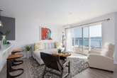 This single-floor residence combines contemporary design with colorful personality, offering a bright and welcoming atmosphere throughout. The open-plan living and dining area serves as the heart of the space, anchored by large sliding glass doors and expansive windows that frame impressive waterfront views, flooding the interior with natural light. A bold teal accent wall defines the dining nook, where a bistro-style table and modern ghost chairs add a playful touch. The living area is generously proportioned, featuring light hardwood-style flooring, a plush sectional sofa, and a striking built-in wall unit with ample storage and display space. A natural wood slab coffee table and vibrant pop-art wall piece add eclectic character to the room. The kitchen is outfitted with white cabinetry, stainless steel appliances including an over-the-range microwave, a mosaic tile backsplash, and clean white countertops. The bedroom is a calm retreat with soft carpeting, a large window with blinds, and colorful striped artwork above the bed. The bathroom features a white vanity, a soaking tub with shower, and warm wood-tone flooring, completing this well-appointed and visually dynamic living space.