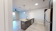 This thoughtfully designed 1-bedroom, 1-bathroom unit offers an efficient and modern single-floor layout. The open-concept kitchen and living area serves as the heart of the unit, anchored by a large granite-topped island with a stainless steel sink and pendant lighting overhead. Dark espresso cabinetry lines the kitchen walls, complemented by a white subway tile backsplash, stainless steel appliances including a refrigerator, range, and microwave, and a built-in dishwasher. Light wood-look flooring flows throughout the main living areas, adding warmth and continuity to the space. The living room is bright and airy, featuring a ceiling fan, neutral walls, and a glass-paneled exterior door that invites natural light and outdoor access. The bedroom is carpeted for comfort, appointed with a ceiling fan, multiple windows with blinds, and direct access to a dedicated walk-in closet equipped with wire shelving on multiple levels for ample storage. The bathroom maintains the consistent design language of the unit, featuring a granite-topped vanity with dark cabinetry, a large mirror, and accessible grab bars. Clean lines, neutral tones, and quality finishes throughout create a cohesive and contemporary living environment.