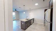 This thoughtfully designed 1-bedroom, 1-bathroom unit offers an efficient and modern single-floor layout. The open-concept kitchen and living area serves as the heart of the unit, anchored by a large granite-topped island with a stainless steel sink and pendant lighting overhead. Dark espresso cabinetry lines the kitchen walls, complemented by a white subway tile backsplash, stainless steel appliances including a refrigerator, range, and microwave, and a built-in dishwasher. Light wood-look flooring flows throughout the main living areas, adding warmth and continuity to the space. The living room is bright and airy, featuring a ceiling fan, neutral walls, and a glass-paneled exterior door that invites natural light and outdoor access. The bedroom is carpeted for comfort, appointed with a ceiling fan, multiple windows with blinds, and direct access to a dedicated walk-in closet equipped with wire shelving on multiple levels for ample storage. The bathroom maintains the consistent design language of the unit, featuring a granite-topped vanity with dark cabinetry, a large mirror, and accessible grab bars. Clean lines, neutral tones, and quality finishes throughout create a cohesive and contemporary living environment.