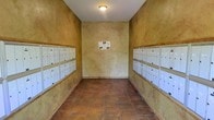 This mail room is a compact, single-floor space designed to serve a multi-unit community. The room is defined by its warm, textured stucco walls rendered in earthy golden and ochre tones, giving the space a distinctly Mediterranean or Southwestern character. The walls on both sides are lined with rows of individual white metal mailboxes, organized in multiple tiers to efficiently accommodate a large number of residents. The mailboxes are uniformly arranged, providing a clean and orderly appearance that makes locating individual units straightforward.

The flooring consists of terracotta-toned ceramic tiles, complementing the warm palette of the stucco walls and reinforcing the overall earthy aesthetic of the space. A single flush-mount ceiling light provides overhead illumination against the white ceiling, keeping the room functional and well-lit. A community notice board is mounted on the far end wall, serving as a central point for shared information and announcements.

The narrow, corridor-like layout of the room is efficient and purposeful, directing foot traffic naturally along the length of the mailboxes on either side. Overall, the space is straightforward in function while maintaining a cohesive, warm architectural character consistent with a Mediterranean-inspired design.