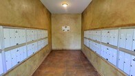 This mail room is a compact, single-floor space designed to serve a multi-unit community. The room is defined by its warm, textured stucco walls rendered in earthy golden and ochre tones, giving the space a distinctly Mediterranean or Southwestern character. The walls on both sides are lined with rows of individual white metal mailboxes, organized in multiple tiers to efficiently accommodate a large number of residents. The mailboxes are uniformly arranged, providing a clean and orderly appearance that makes locating individual units straightforward.

The flooring consists of terracotta-toned ceramic tiles, complementing the warm palette of the stucco walls and reinforcing the overall earthy aesthetic of the space. A single flush-mount ceiling light provides overhead illumination against the white ceiling, keeping the room functional and well-lit. A community notice board is mounted on the far end wall, serving as a central point for shared information and announcements.

The narrow, corridor-like layout of the room is efficient and purposeful, directing foot traffic naturally along the length of the mailboxes on either side. Overall, the space is straightforward in function while maintaining a cohesive, warm architectural character consistent with a Mediterranean-inspired design.