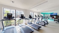 This fitness center on the lower level offers a comprehensive workout environment designed to support a wide range of exercise routines. The space is bright and open, with large windows along one wall that flood the cardio area with natural light and provide views of the surrounding outdoor space. The clean, white walls are accented by bold teal feature walls, adding a vibrant and energizing atmosphere throughout.

The cardio section is well-appointed with a row of treadmills and elliptical machines, all equipped with individual digital consoles. Adjacent to the cardio area, a dedicated functional training zone features a multi-station cable machine, a punching bag, suspension training equipment, plyo boxes, and a large wall mirror that enhances the sense of space. This area benefits from soaring high ceilings, making it ideal for dynamic, full-range movements.

A staircase connects the lower level to additional areas, and a cable crossover machine is positioned near the base of the stairs, maximizing use of the available floor space. The flooring throughout is a durable, light-toned rubberized surface suited for both cardio and strength activities. Additional strength and resistance training machines are visible toward the back of the space. Overall, the fitness center is thoughtfully laid out to accommodate diverse training needs in a modern, well-lit setting.