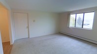 This single-floor space presents a clean, open living area characterized by neutral cream-colored walls and light-toned wall-to-wall carpeting that contributes to a calm, understated ambiance. The room offers a generous sense of openness, with sufficient space for a variety of furniture arrangements. A window along one wall allows natural light to filter into the interior, offering a view of surrounding greenery outside. Baseboard heating runs along the base of the exterior wall, providing an efficient and unobtrusive heating solution. A standard entry door is positioned on one side of the room, featuring a simple, functional design consistent with the overall aesthetic of the space. A wall-mounted panel, likely a thermostat or intercom unit, is visible near the entry. The plain white ceiling complements the light palette of the room, keeping the atmosphere bright and neutral. Overall, the space is straightforward and well-maintained, offering a blank canvas that can be easily personalized or furnished to suit a range of needs and preferences.