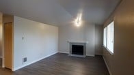 This single-floor space offers a clean, modern aesthetic with a welcoming and functional layout. The living area serves as the centerpiece of the space, featuring rich, dark hardwood-style flooring that runs throughout, creating a warm and cohesive look. Crisp, neutral-toned walls provide a versatile backdrop that complements a wide range of décor styles.

A standout feature of the living area is the classic fireplace, framed by a light-toned tile surround and a simple white mantel, offering both a decorative focal point and a source of warmth. The angled, vaulted ceiling adds a sense of height and openness to the room, enhancing the overall sense of space. A multi-bulb ceiling fixture provides direct overhead lighting, while a window along the side wall admits natural light, brightening the interior.

The clean lines of the baseboards and the smooth, textured ceiling finish contribute to the well-maintained and move-in-ready feel of the space. The open, uncluttered layout offers flexibility for a variety of furniture arrangements, making the living area both practical and inviting. Overall, this space balances simplicity with comfort, making it an ideal setting for everyday living.