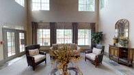 This elegantly appointed single-floor community space makes a strong first impression with its soaring ceilings and open, airy layout. The grand lobby area features slate tile flooring, classic white columns, and recessed lighting that work together to create a bright and inviting atmosphere. Generous windows — including clerestory windows set high on the walls — flood the interior with abundant natural light while offering views of the surrounding outdoor greenery.

A standout focal point is the beautifully crafted fireplace, framed by a white stone mantle and accented with a decorative slate tile surround and an ornate gold-toned fire screen. Arched mirrors and curated accessories on nearby consoles add a refined, classic touch to the space.

Seating areas throughout are thoughtfully arranged with upholstered wingback chairs, chaise lounges, and coordinating accent furnishings, providing comfortable spaces for conversation and relaxation. A dedicated bar area, clad in stacked stone and finished with a dark countertop and bar stools, offers a warm, rustic contrast to the polished surroundings. French doors lead directly to an outdoor patio, extending the living space further. The overall ambiance is sophisticated yet welcoming, combining classic architectural details with warm, layered décor.