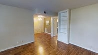 This single-story space showcases warm hardwood flooring that flows consistently throughout, creating a cohesive and inviting atmosphere. The main living area is open and airy, benefiting from a clean, neutral color palette of white trim and soft beige walls that maximize the sense of space. A standout feature is the built-in white shelving unit positioned along one wall, offering both display and storage opportunities while adding an architectural detail to the room.

An arched passageway connects the living area to the dining space beyond, where an elegant crystal chandelier serves as a focal point, casting warm ambient light across the room. The dining area's warm-toned walls complement the rich hues of the hardwood flooring, contributing to an overall sense of warmth and intimacy.

Additional doorways visible from the main area suggest further rooms beyond, providing a functional layout throughout the home. White baseboards and trim throughout provide a crisp contrast to the warm flooring and walls. The space overall presents a well-maintained interior that balances classic decorative touches with practical, everyday livability.