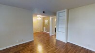 This single-story space showcases warm hardwood flooring that flows consistently throughout, creating a cohesive and inviting atmosphere. The main living area is open and airy, benefiting from a clean, neutral color palette of white trim and soft beige walls that maximize the sense of space. A standout feature is the built-in white shelving unit positioned along one wall, offering both display and storage opportunities while adding an architectural detail to the room.

An arched passageway connects the living area to the dining space beyond, where an elegant crystal chandelier serves as a focal point, casting warm ambient light across the room. The dining area's warm-toned walls complement the rich hues of the hardwood flooring, contributing to an overall sense of warmth and intimacy.

Additional doorways visible from the main area suggest further rooms beyond, providing a functional layout throughout the home. White baseboards and trim throughout provide a crisp contrast to the warm flooring and walls. The space overall presents a well-maintained interior that balances classic decorative touches with practical, everyday livability.