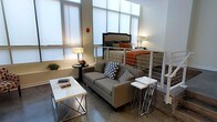 This single-floor space showcases a clever split-level layout, where an elevated sleeping loft overlooks the main living area below, separated by a sleek white cable railing system. The loft accommodates a full-sized bed with an upholstered headboard and warm-toned bedding, creating an intimate retreat above the open living space.

The main level features polished concrete floors that lend an industrial character to the interior. A gray upholstered sofa anchors the living area, complemented by a modern white coffee table with chrome cross-leg detailing and a matching set of nesting side tables. A small round side table with a table lamp adds warmth and a finishing touch to the seating arrangement.

Large floor-to-ceiling windows dressed with roller shades flood the space with abundant natural light, enhancing the airy, open feel of the interior. The white walls and clean architectural lines contribute to a contemporary aesthetic throughout. A staircase with white framing provides access to the loft level. Overall, the space balances industrial elements with modern furnishings to create a functional and stylish environment.