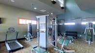 This fitness center offers a comprehensive workout environment designed to accommodate a range of exercise routines. The space features soaring ceilings that create an open, airy atmosphere, enhanced by natural light streaming in through shuttered windows positioned along the walls. Recessed lighting supplements the natural illumination, ensuring the space remains bright and well-lit throughout.

The room is outfitted with a multi-station cable and resistance training machine at its center, serving as a focal point for strength training. Surrounding it are additional weight machines, adjustable benches, and a well-stocked dumbbell rack offering a graduated selection of free weights. The layout is thoughtfully arranged to allow users to move comfortably between stations without congestion.

Wall-mounted mirrors are positioned to allow users to monitor their form during workouts, contributing to both the functionality and the sense of depth within the space. The flooring features a durable, patterned surface suited to the demands of a fitness environment. Subtle decorative touches, including wall sconces and an accent color band near the ceiling, add a polished finish to this practical and well-appointed facility.