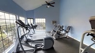 This single-floor fitness center offers a focused and functional workout environment designed to support a range of cardio-based exercise routines. The room is finished with light blue walls that create a calm, motivating atmosphere, complemented by dark rubberized flooring that provides both durability and traction underfoot.

The space is equipped with a selection of cardio machines, including treadmills, an elliptical trainer by Precor, an upright exercise bike, and a recumbent bike — offering users a variety of low- and high-impact workout options. The equipment is arranged to allow comfortable use and circulation throughout the room.

Architecturally, the room features vaulted ceilings that lend a sense of openness and airiness to the space. A ceiling fan is centrally mounted, providing continuous air circulation during workouts. Multiple windows along one wall allow natural light to filter in, creating a bright and energizing environment while offering views of the outdoor greenery beyond. A wall-mounted air conditioning unit provides additional climate control for year-round comfort. The textured ceiling adds a subtle finishing detail to the overall aesthetic. Together, these elements make this fitness center a well-appointed and practical space for daily exercise.