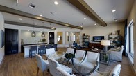 This single-story space offers an open-concept layout that seamlessly integrates multiple functional areas into one cohesive environment. The interior is characterized by neutral white walls, wood-look tile flooring, and a ceiling accented with dark exposed beams that add architectural depth and visual interest.

The lounge area serves as the focal point of the space, furnished with elegant white upholstered armchairs, blue-and-white striped accent chairs, and a glass-top coffee table, creating a comfortable and sophisticated seating arrangement. Recessed lighting throughout the ceiling provides consistent illumination, complemented by stylish pendant lights that hang above the kitchen bar area.

The kitchen bar features a granite countertop, bar-height seating, and stainless steel appliances, making it a functional and attractive gathering spot. A media area with a wall-mounted flat-screen television and an open shelving unit adds entertainment functionality to the space.

Natural light filters in through multiple glass doors and windows, enhancing the bright, airy ambiance. The overall design balances modern aesthetics with comfortable, welcoming furnishings, creating a versatile and inviting communal environment.