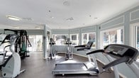 This fitness center offers a bright and welcoming environment designed to support an active lifestyle. The space is arranged on a single floor and is defined by its curved wall of large windows that wrap around the room, flooding the interior with natural light and providing open views of the surrounding greenery outdoors. The soft, muted blue-gray wall paneling accented with crisp white trim gives the space a clean, calming aesthetic that complements its purpose. Recessed lighting and additional ceiling fixtures ensure the room remains well-lit at all hours. The floor is covered in durable, textured rubber matting suited for high-traffic fitness use. The cardio area is thoughtfully laid out with multiple treadmills, elliptical trainers, and other exercise machines, all positioned to take advantage of the natural light and outdoor views. The textured ceiling adds a subtle design element overhead, while ventilation is well integrated throughout. The overall ambiance is airy and motivating, striking a balance between functionality and a pleasant, light-filled atmosphere that makes for a comfortable workout environment.