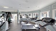 This fitness center offers a bright and welcoming environment designed to support an active lifestyle. The space is arranged on a single floor and is defined by its curved wall of large windows that wrap around the room, flooding the interior with natural light and providing open views of the surrounding greenery outdoors. The soft, muted blue-gray wall paneling accented with crisp white trim gives the space a clean, calming aesthetic that complements its purpose. Recessed lighting and additional ceiling fixtures ensure the room remains well-lit at all hours. The floor is covered in durable, textured rubber matting suited for high-traffic fitness use. The cardio area is thoughtfully laid out with multiple treadmills, elliptical trainers, and other exercise machines, all positioned to take advantage of the natural light and outdoor views. The textured ceiling adds a subtle design element overhead, while ventilation is well integrated throughout. The overall ambiance is airy and motivating, striking a balance between functionality and a pleasant, light-filled atmosphere that makes for a comfortable workout environment.