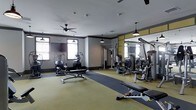 This single-floor fitness center is organized into two distinct functional areas. The main workout zone features resilient yellow-toned flooring with a central dark rubber mat runner, providing designated space for both machine-based training and floor exercises. The room is furnished with a comprehensive selection of equipment, including elliptical machines, rowing machines, cable and multi-use strength machines, and adjustable benches, accommodating a wide range of fitness routines. Mirrored wall panels with gold-toned accent trim run along one side, visually expanding the space while allowing users to monitor their form. Ceiling fans and pendant lighting fixtures are distributed throughout, ensuring comfortable air circulation and ample illumination. Windows along the perimeter walls bring in natural light and contribute to an open, airy atmosphere. The secondary area, separated by a partial partition wall, offers an open-plan space suited for stretching, yoga, or functional training. Gray accent walls with white trim give the space a clean, modern aesthetic throughout.
