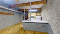 This one-bedroom, one-bathroom unit offers a distinctive industrial aesthetic defined by its exposed corrugated metal ceiling, visible ductwork, and raw, textured concrete-style walls. Warm hardwood floors run throughout the entire space, providing a natural contrast to the rugged architectural elements above.

The open living area greets residents at entry through a bold blue front door, complemented by a ceiling fan and a window fitted with blinds that allows natural light to filter in. The textured walls and exposed wood beams give the living space a loft-like character.

The kitchen is efficiently laid out and accessible from the living area via a peninsula counter with a double stainless steel sink. Light wood cabinetry lines the walls, offering ample storage, while white appliances — including a freestanding electric range and full-size refrigerator — provide all essential cooking amenities. Exposed metal support columns add to the industrial feel.

The bedroom is a well-proportioned retreat that continues the same hardwood flooring and textured wall aesthetic, with exposed ductwork and a ceiling fan overhead providing both function and style.

The bathroom is cleanly appointed, featuring a tiled shower-tub combo, toilet, and an open wood-framed storage shelf, finished with tile flooring. The overall ambiance of this space is utilitarian yet characterful, ideal for those who appreciate raw, industrial design.